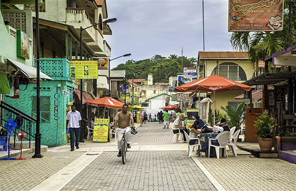 A street scene with people walking and sitting at outdoor tables, colorful buildings on either side, and a person riding a bicycle down the center.