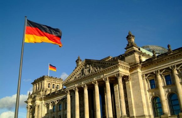 A German flag waves in front of a classical building, possibly governmental, under a lightly clouded blue sky.