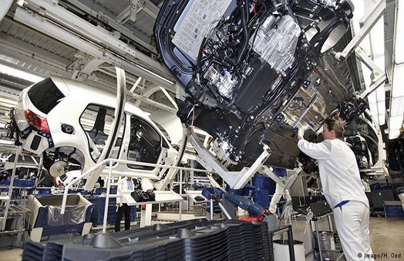 A worker in a white uniform is assembling a car at an automotive manufacturing plant, with the car chassis lifted above the ground.