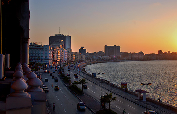 A cityscape during sunset with buildings along a waterfront, street lights illuminated, and a colorful sky transitioning from blue to orange hues.