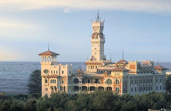 A historic ornate building with a prominent tower, situated near a coastline with trees in the foreground and a clear sky in the background.