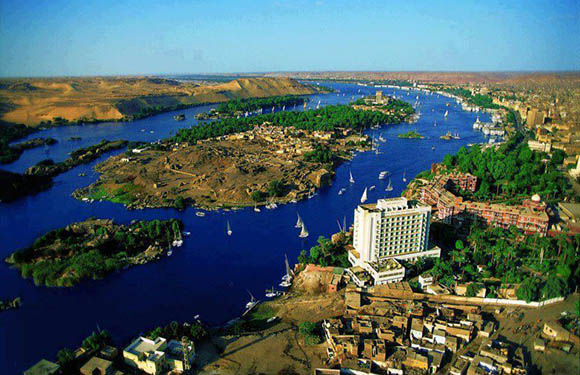 Aerial view of a wide river with scattered islands, traditional sailboats, and buildings along the banks under a clear blue sky.