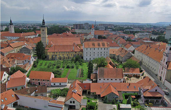 Aerial view of a historic town with dense buildings, featuring a prominent church tower, red-tiled roofs, and green spaces amidst an urban layout.