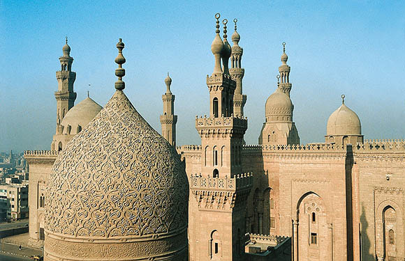 Aerial view of an ornate mosque with multiple minarets and a large dome, set against a clear sky in an urban environment.