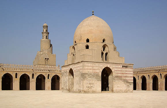 A historical stone building with a large central dome and smaller surrounding structures, set against a clear blue sky.