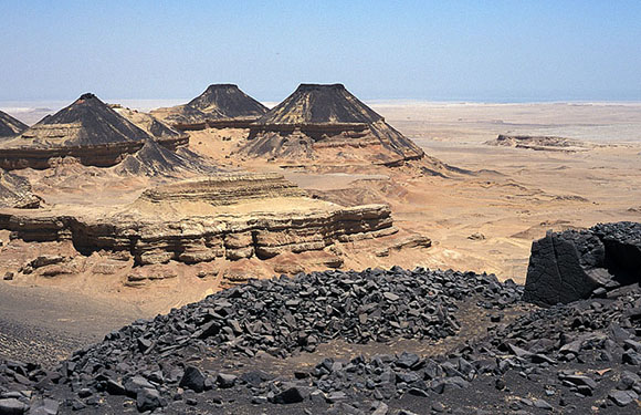 A landscape featuring several dark, conical hills rising from a flat, arid desert plain under a clear sky.