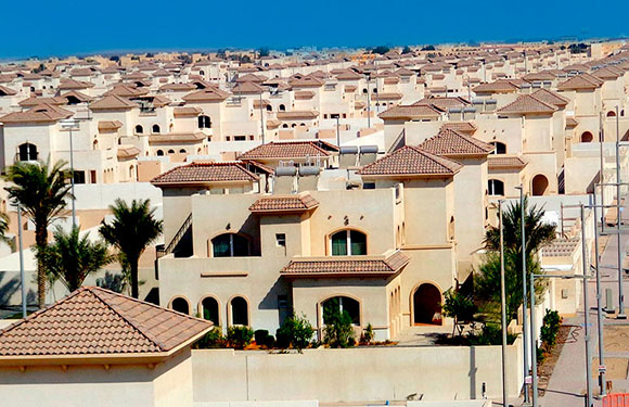 A dense cluster of similar-looking, beige-colored houses with terracotta roofs, closely packed in a suburban development.