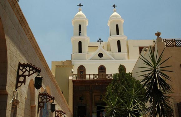 A church with two white bell towers against a clear sky, surrounded by buildings with Middle Eastern architectural features.