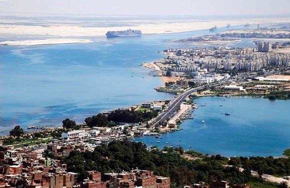 Aerial view of a coastal cityscape with a road running through it, adjacent to a body of water, with a ship visible in the distance.