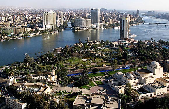 Aerial view of a city with buildings along the banks of a wide river, greenery, and clear skies.