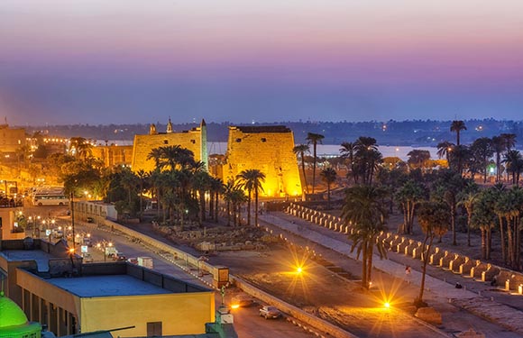 An image of a historic fortress with illuminated walls at dusk, palm trees lining the path, and a soft purple and orange sky in the background.