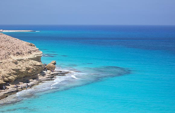 A coastal landscape with clear turquoise waters, a rocky shoreline on the left, and a clear blue sky above.