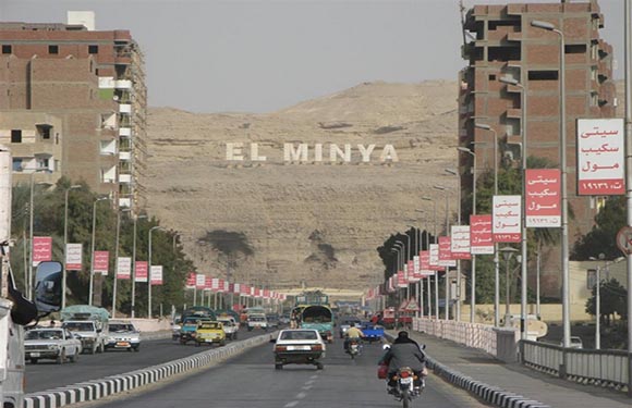 A street view with vehicles and buildings on either side, and a hill in the background with "EL MINYA" written on it.