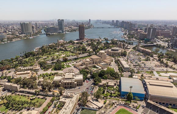 Aerial view of a densely built urban area with numerous buildings surrounded by a wide river, with a clear sky above.