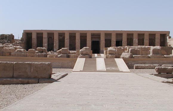 An image of an ancient temple with columns, showing the front facade and entrance, with clear skies above and ruins in the foreground.