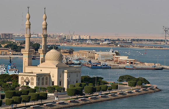 A coastal cityscape features a mosque with two minarets and a dome, overlooking a sea with boats and a distant skyline.