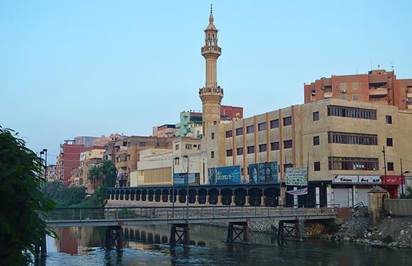 A cityscape with buildings along a river, featuring a prominent minaret rising above the structures under a clear sky.