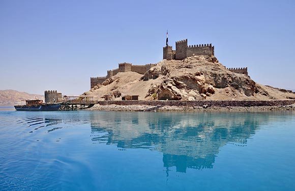 A castle on a hill with a clear blue sky and calm blue water in the foreground.