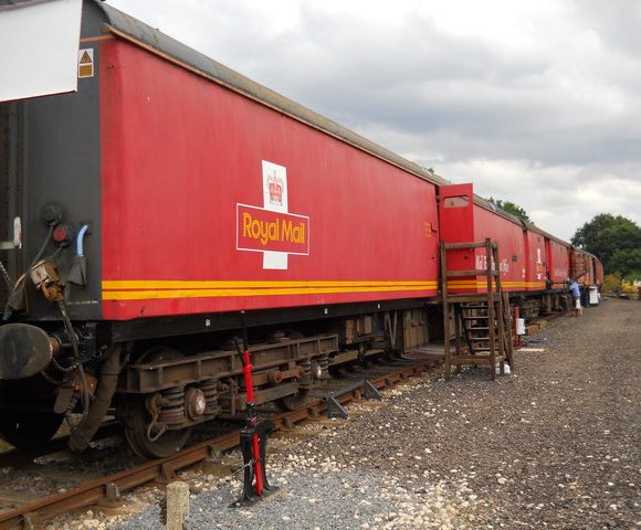 A row of red Royal Mail train carriages on railway tracks with a cloudy sky in the background.