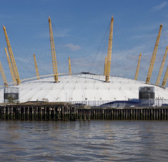 A photo shows a large dome building with a white roof and yellow supports, located near water under a clear sky.