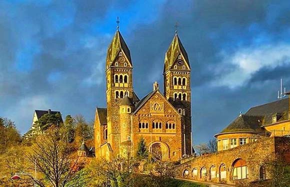 An image of a Romanesque church with twin spires against a blue sky with clouds. Trees and greenery are visible around the building.