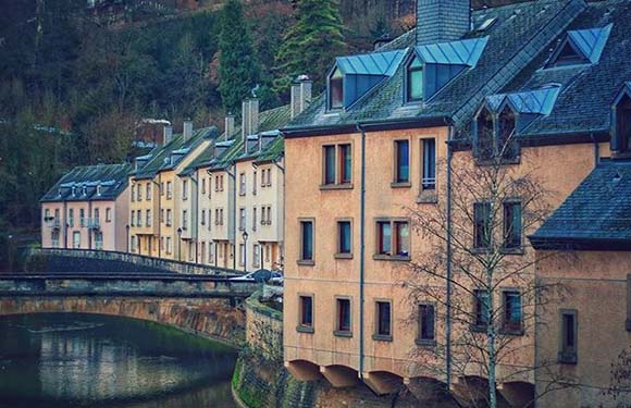 Colorful European buildings line a calm river, with a bridge and trees partially hidden in the background.