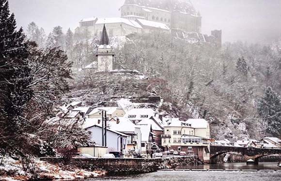 A snowy scene featuring a river, riverside buildings, a bridge, and a hilltop castle under a gray sky.