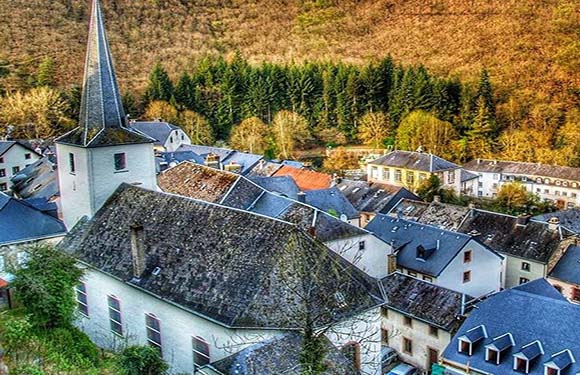 A scenic European village with traditional homes and a church, nestled in a valley amid forested hills.