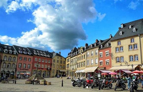 A colorful town square with historic buildings, outdoor seating at cafes, motorcycles parked in the foreground, and a blue sky with clouds overhead.