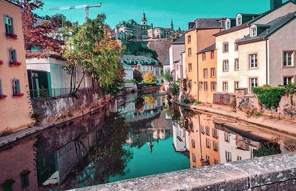 A picturesque view of a calm river flowing through a quaint town with colorful buildings on either side and greenery in the background.