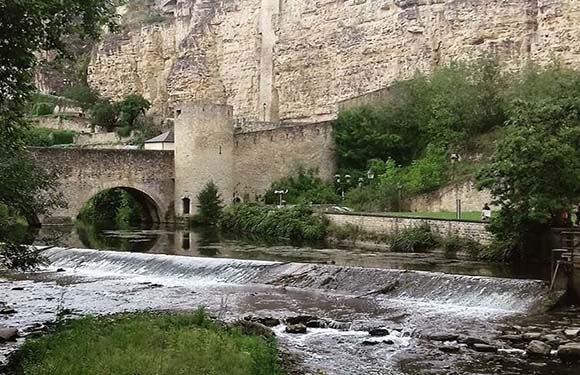 A stone bridge arches over a calm river with a waterfall, encircled by greenery and a tall cliff.