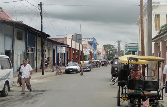 A bustling street scene with pedestrians, vehicles, vibrant buildings, a bicycle taxi in the foreground, under a cloudy sky.