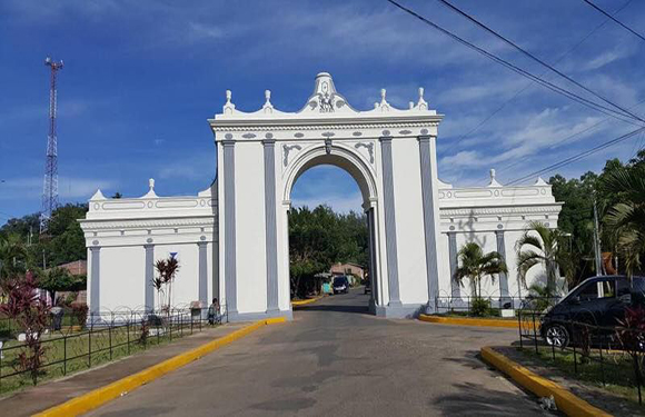 A white, intricately designed archway marks the entrance of a park, with trees, a road, and part of a car visible.