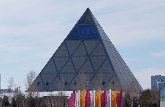 A pyramid-shaped building with a geometric facade under a partly cloudy sky, with colorful flags in the foreground.