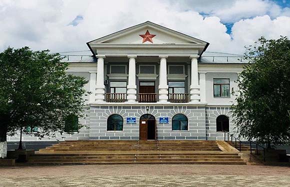 A two-story white building with a central entrance, columns, a red star at the peak, and a front staircase under a cloudy sky.