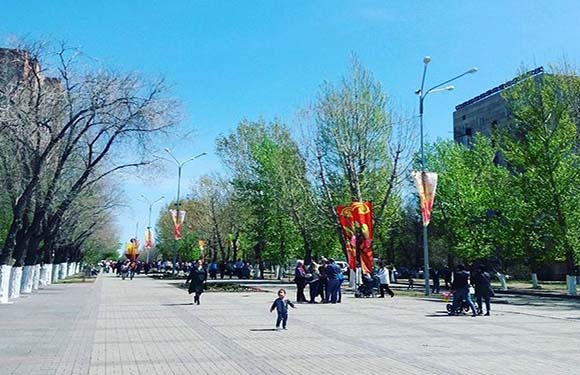 A photo shows people strolling on a tree-lined promenade with festive banners and buildings under a clear sky.