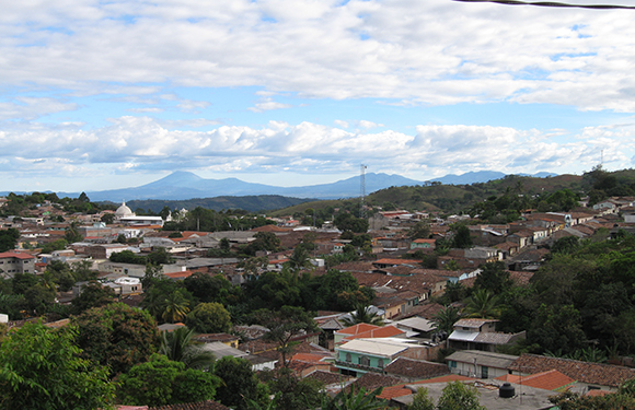 A panoramic view of a town with dense housing and trees, under a partly cloudy sky with mountains in the distance.