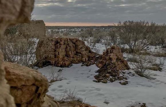 An image showing a snowy landscape at dusk with ruins of stone structures in the foreground and bare trees in the background under a cloudy sky.