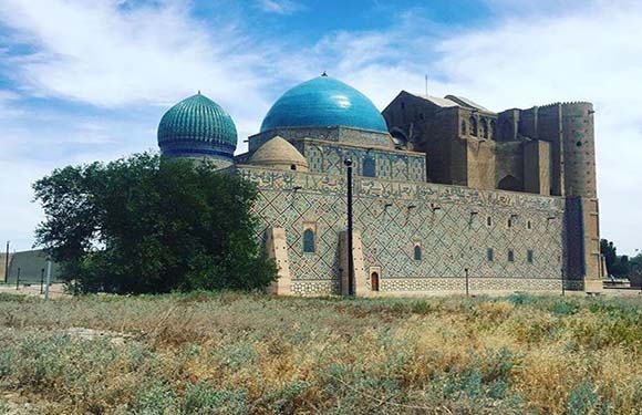 A historical building with a large dome and ornate stone walls, surrounded by a grassy field under a clear sky.