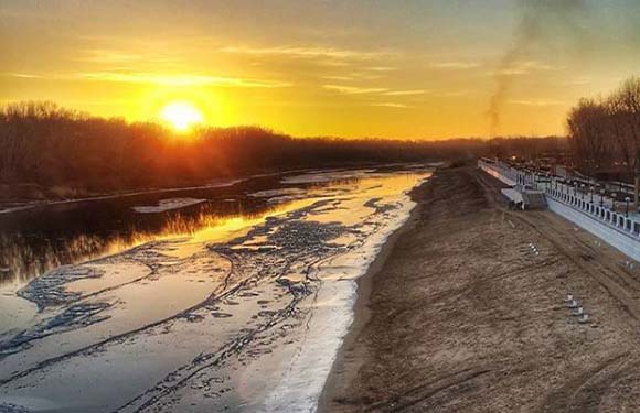 A sunset view over a partially frozen river with trees on the banks and a clear sky.