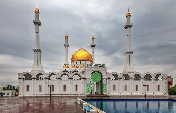 A mosque with white walls and a golden dome flanked by four minarets against a cloudy sky, with a reflective pool in the foreground.