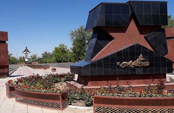 A large black star monument with a golden emblem in the center, surrounded by flower beds and smaller structures, under a clear blue sky.