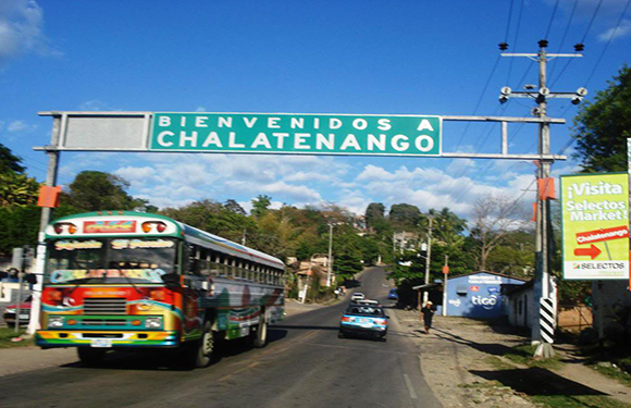 A vibrant bus and car are under a large sign saying "Bienvenidos a Chalatenango", with a scenic backdrop.