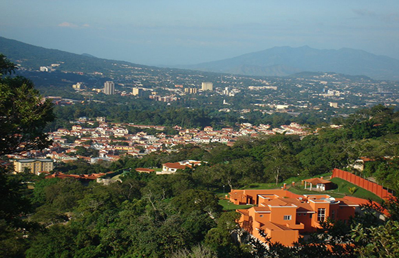 A panoramic view of a city with buildings spread out among green areas, with mountains in the background and a clear sky above.