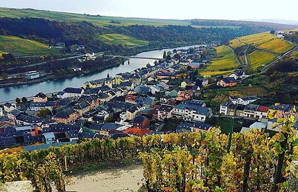 Aerial view of a quaint village with traditional houses alongside a winding river, surrounded by lush hills with vineyards under a partly cloudy sky.