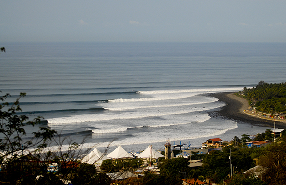 A scenic view of a coastal area with waves rolling onto a curved beach, surrounded by lush greenery and a few buildings.