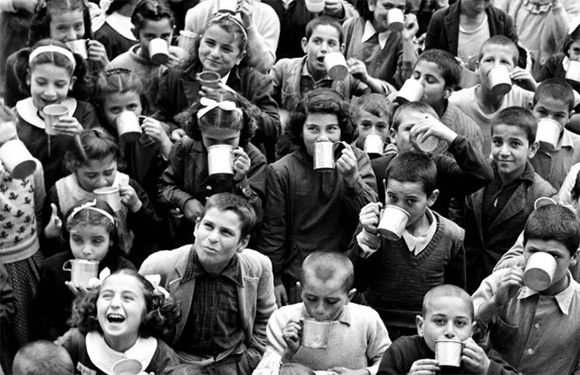 A black and white photo of a group of children of various ages, some holding cups, with a few adults among them, all appearing to be outdoors.