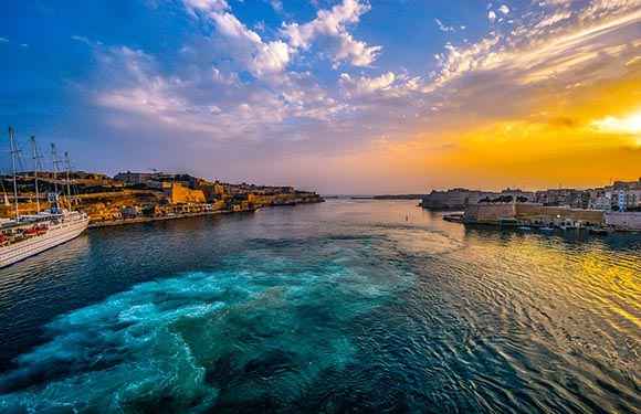 A scenic view of a waterway at sunset with vibrant blue and orange skies, featuring boats docked along the shore and buildings lining the coast.