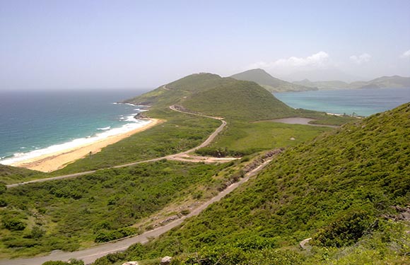 A coastal landscape with a winding road leading through green hills towards a beach with blue waters on one side and a rougher sea on the other.