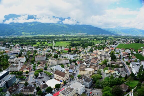 Aerial view of a small town amid green fields and trees, with cloud-covered mountains in the background.